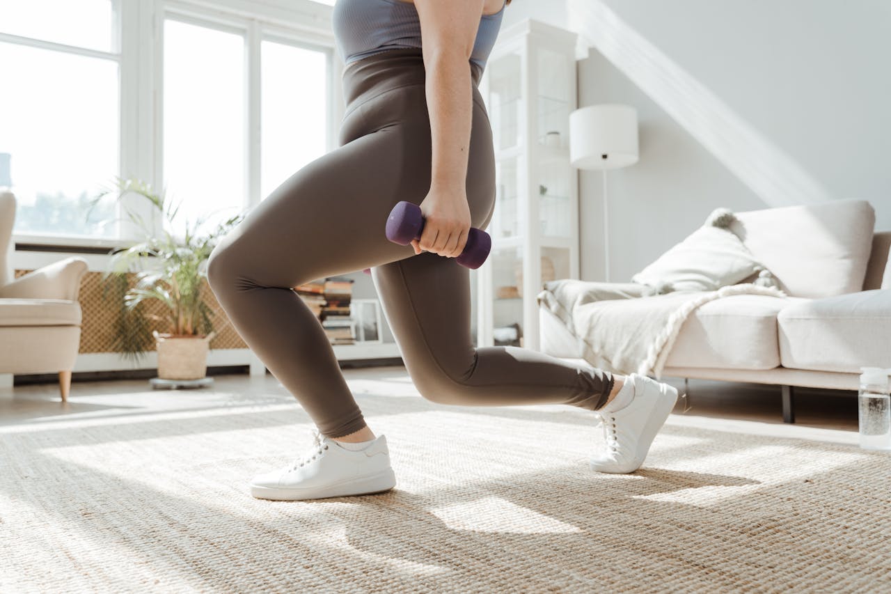 A woman in active wear performs a lunges exercise with dumbbells indoors.